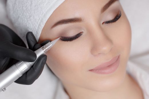 Close up of woman lying on couch at beauty salon with closed eyes and enjoying when cosmetologist making eyeliner permanent make up. Professional wearing black gloves and holding speciality tool.